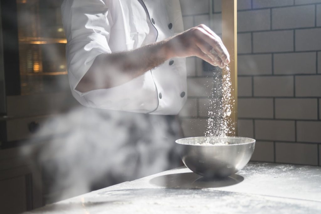 Chef in white uniform sprinkling flour into a steel bowl in a modern kitchen.