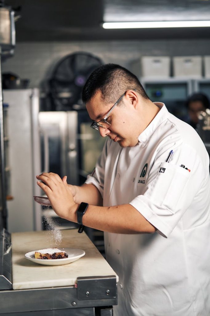 A skilled chef garnishes a dessert with powdered sugar in a modern kitchen setting.